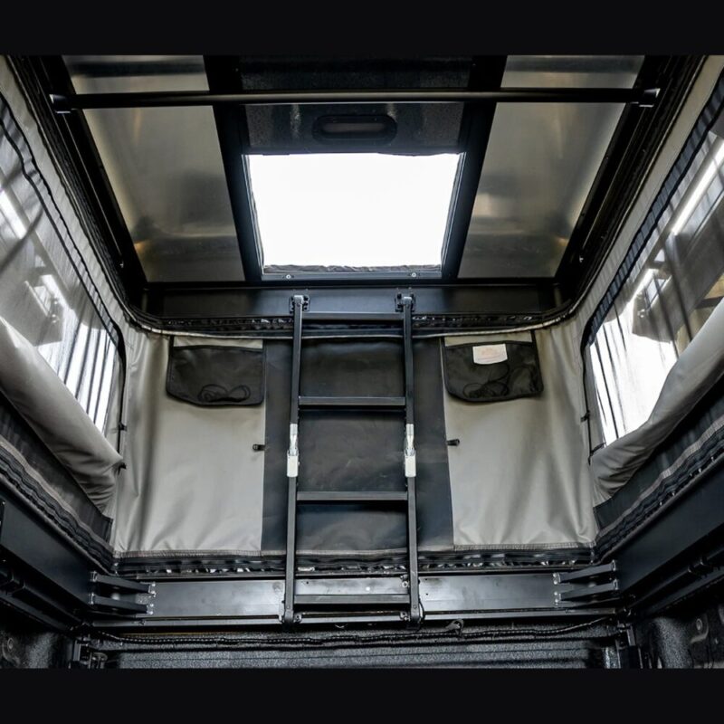 The interior of the Safari Cruiser upon entering from the truck bed. Focus on the ladder leading to the second floor of the two-story tent, through the hatch supported by strong bars, with windows also present on the first floor.