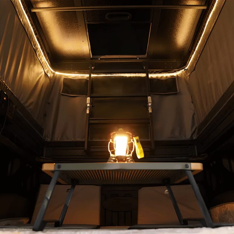 Interior view of the Safari Cruiser Two-Story Car Roof Top Tent by WildLand, featuring an inner ladder, ambient LED lighting, and a lantern on a small table, highlighting its spacious and cozy design for two or three people.